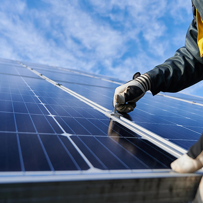 Gloved hand works on a photovoltaic panel, with a clear sky and scattered clouds in the background