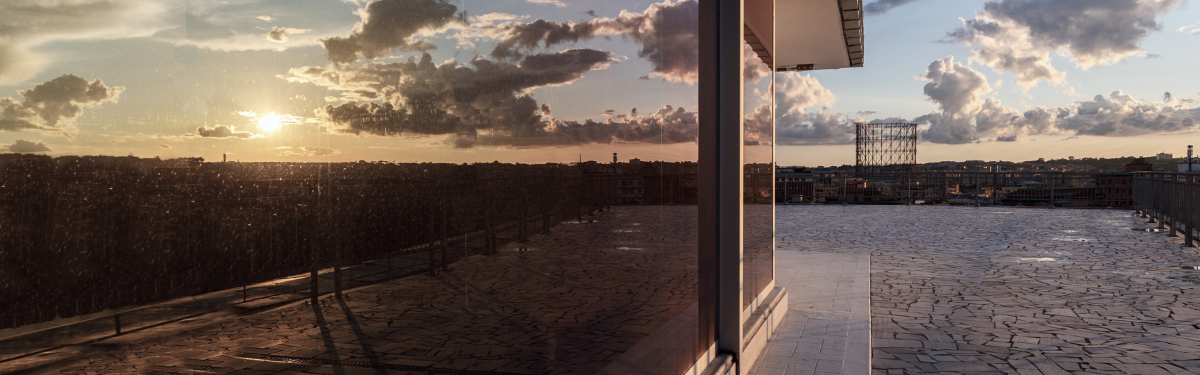 Dettaglio della terrazza della sede del Gruppo Acea al tramonto, con il sole riflesso sulla vetrata sulla sinistra, e con vista sulla città di Roma e il Gazometro sullo sfondo.