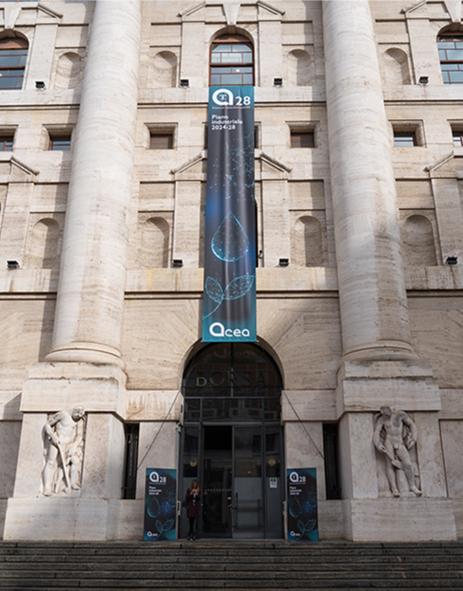 Acea Capital Market Days Ingresso di un grande edificio con alte colonne e statue ai lati della porta. Un banner verticale tra le colonne sopra l'ingresso mostra il logo di Acea e il testo "28" in alto, seguito da "Piano Industriale 2024-2028" e un logo con "Acea" in basso.