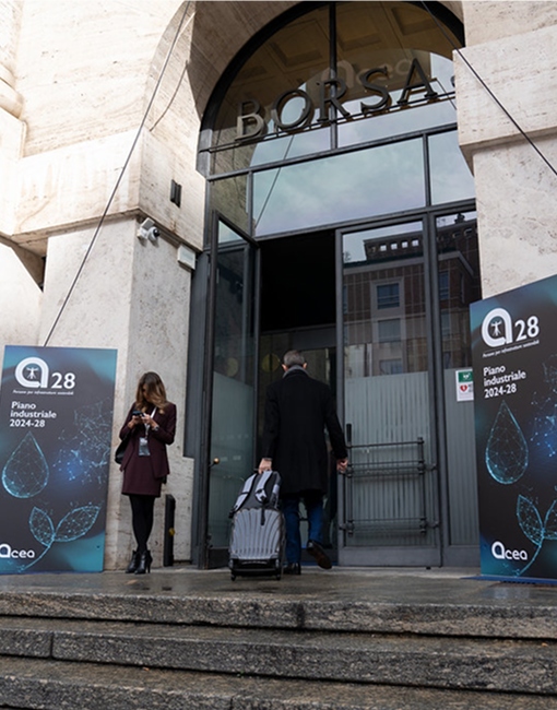 Due persone sono davanti all'ingresso di un edificio: una guarda il telefono, mentre l'altra cammina verso la porta trascinando una valigia. Ai lati dell'ingresso ci sono due grandi banner con il testo "Acea 28 Piano Industriale 2024-28".