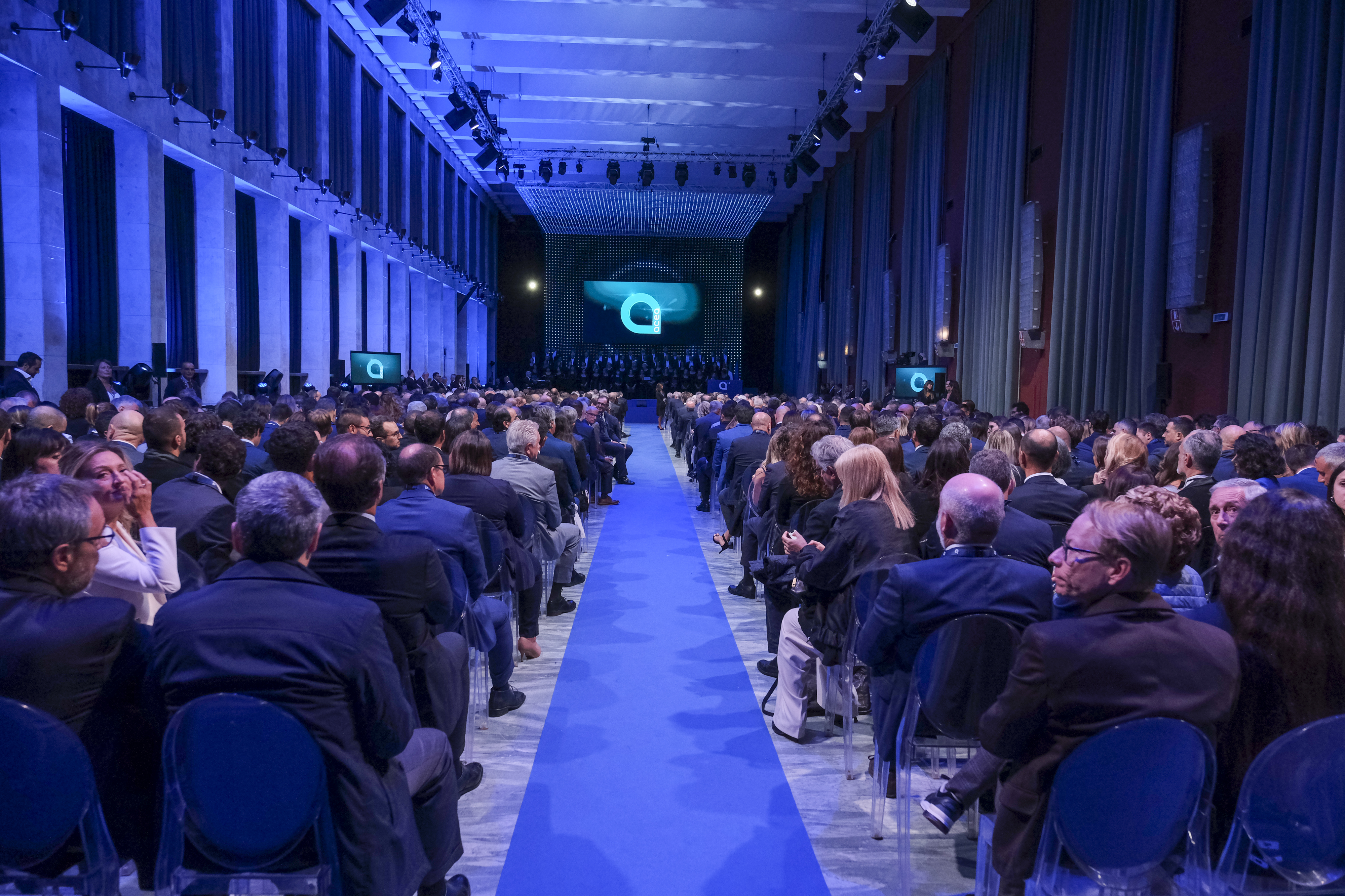 an audience of seated people divided into two aisles; in the centre a blue carpet and in the background a screen with the acea logo projected
