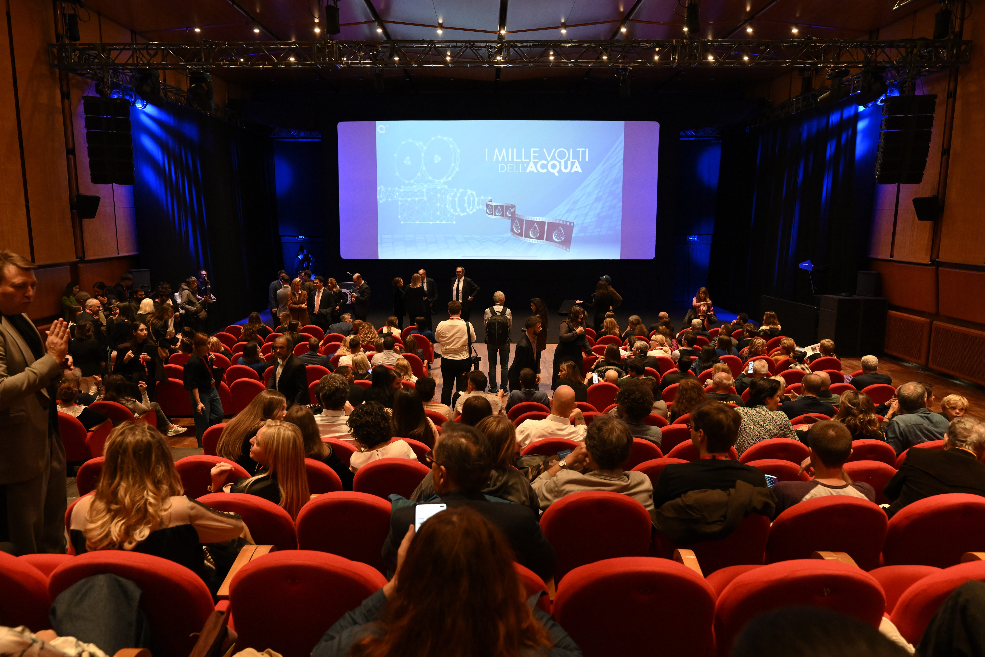 Sala piena di gente e sullo sfondo uno schermo acceso con la proiezione de "i mille volti dell'acqua"