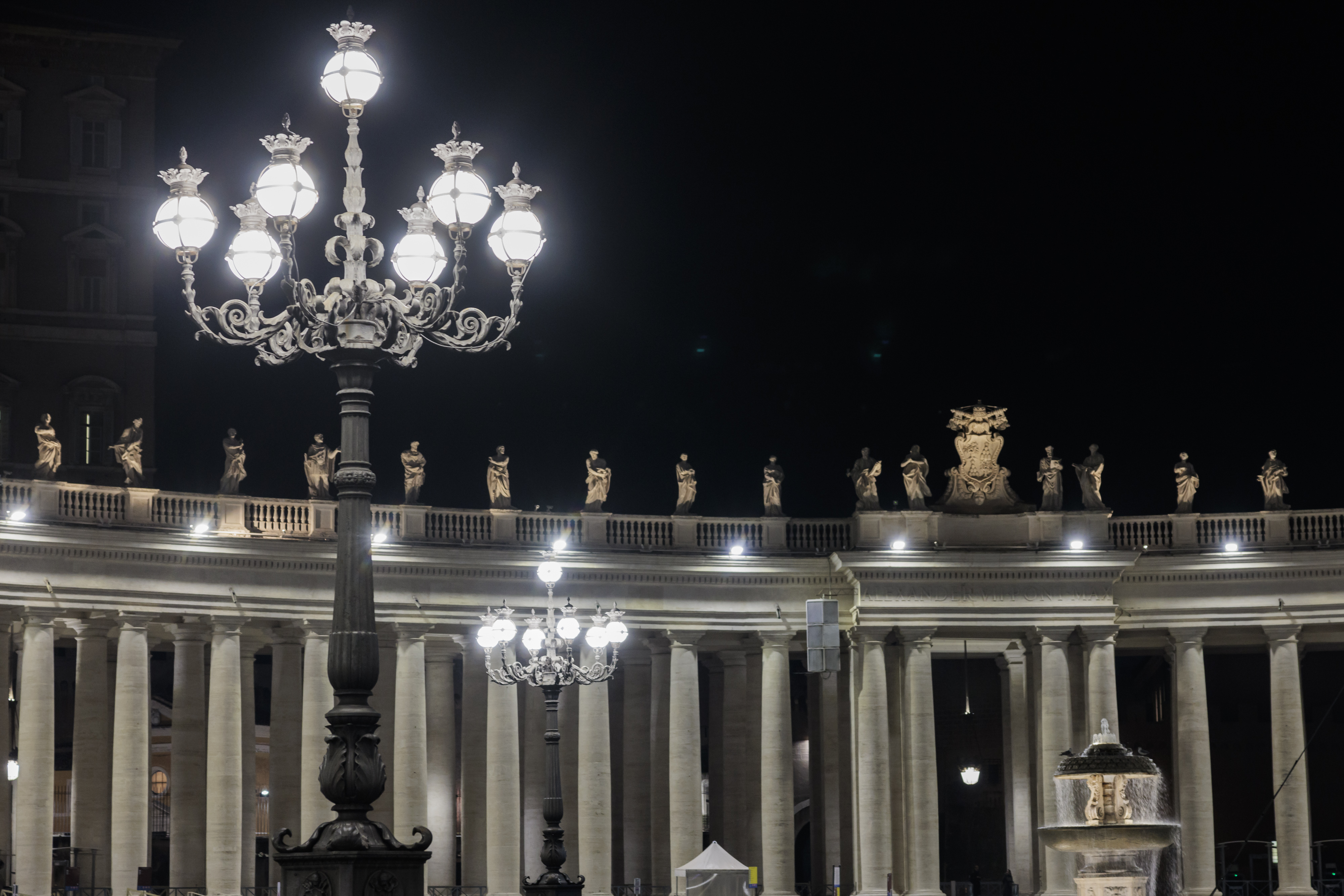 A lit street lamp in the foreground; in the background you can see the illuminated colonnade of St. Peter's Square